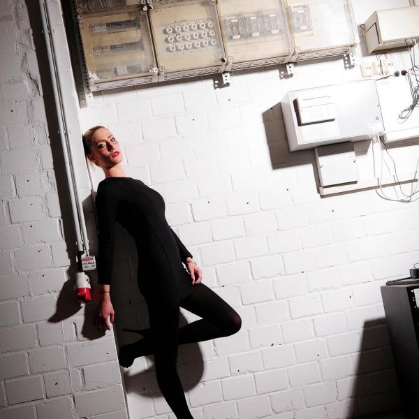 Portraitfoto aus einem Fotoshooting on Location eines weiblichen Models mit blonden Haaren, in einem schwarzen Abendkleid, stehend vor einer weißen Brick Wall mit elektrischen Geräten